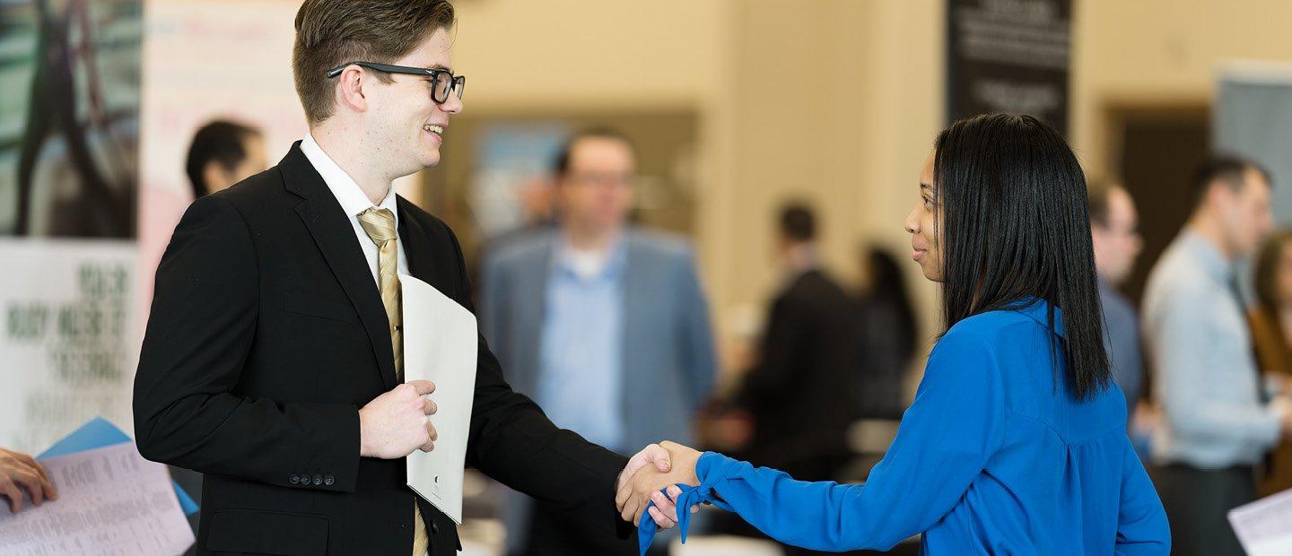 A young man in a suit holding a document, shaking a young woman's hand.