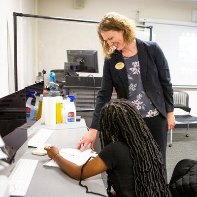 A smiling woman talks to a student seated at a table.