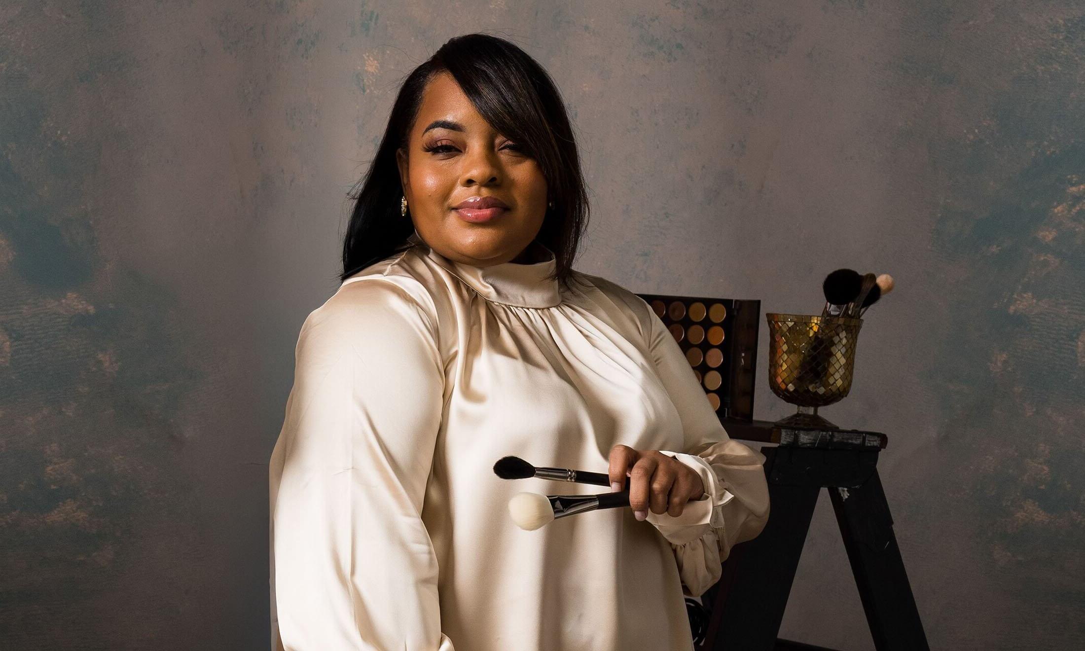 A woman holding makeup brushes looks at the camera.